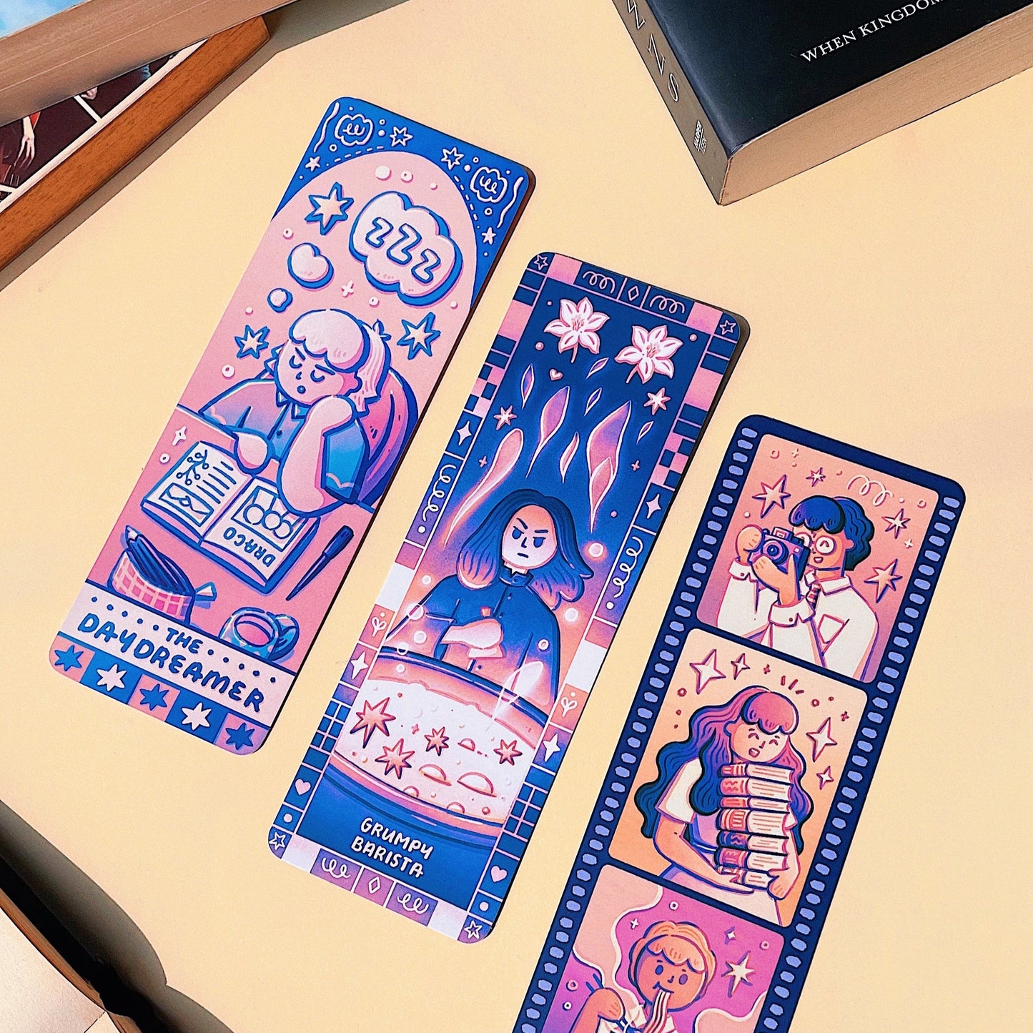 Three colorful bookmarks with illustrations on a beige surface with books in the background.