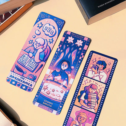Three colorful bookmarks with illustrations on a beige surface with books in the background.